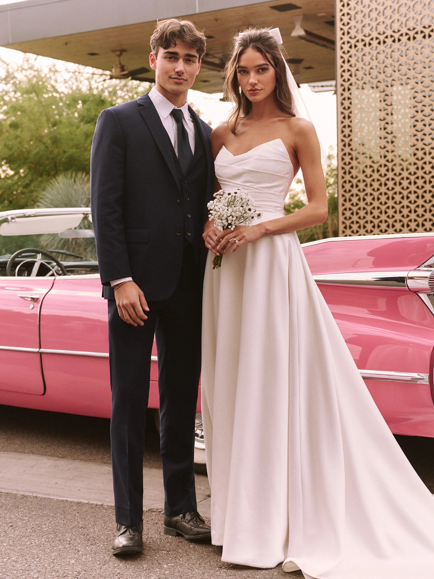 A bride and groom stand in front of a vintage pink Cadillac. The bride is wearing a wedding dress called Ethelinda by Rebecca Ingram and holding a bouquet of flowers, while the groom is dressed in a black suit.